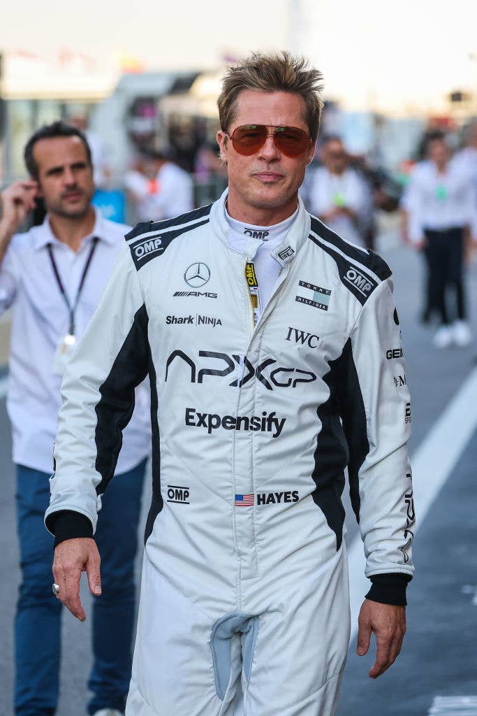 Brad Pitt, wearing a professional racing suit with sponsor logos, walking on a racetrack path