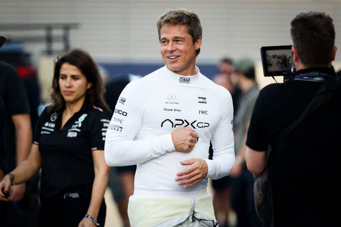 Brad Pitt in a racing suit at an event, with a crowd and camera crew in the background