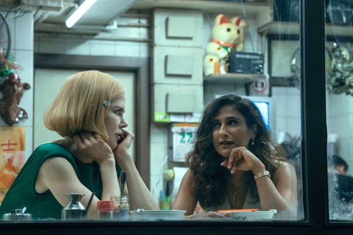 Two women sit at a diner table engaged in conversation, with a maneki-neko cat figure in the background