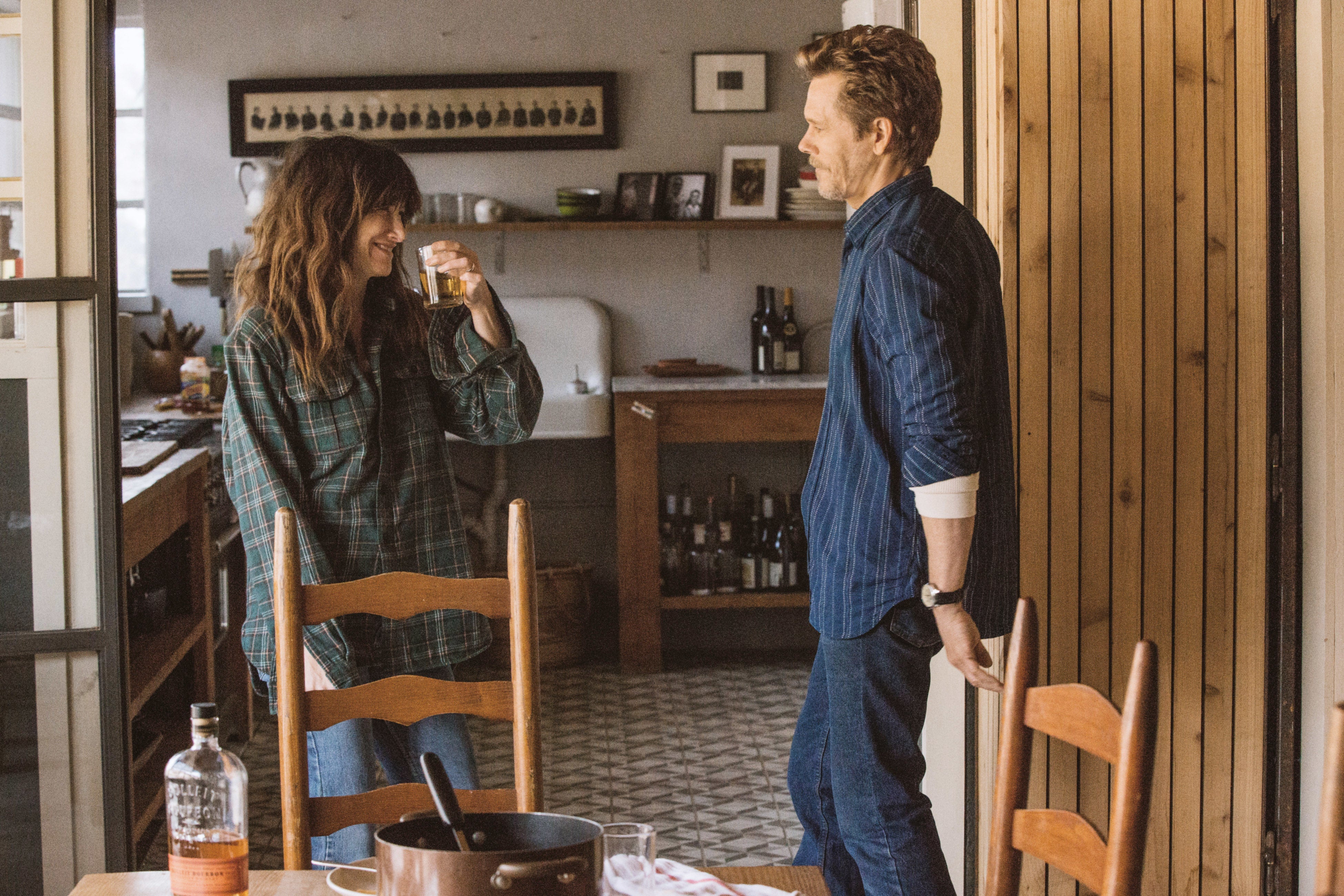 Two people in a kitchen, one holding a drink, casually dressed. They seem engaged in a relaxed conversation with a cozy, rustic setting around them