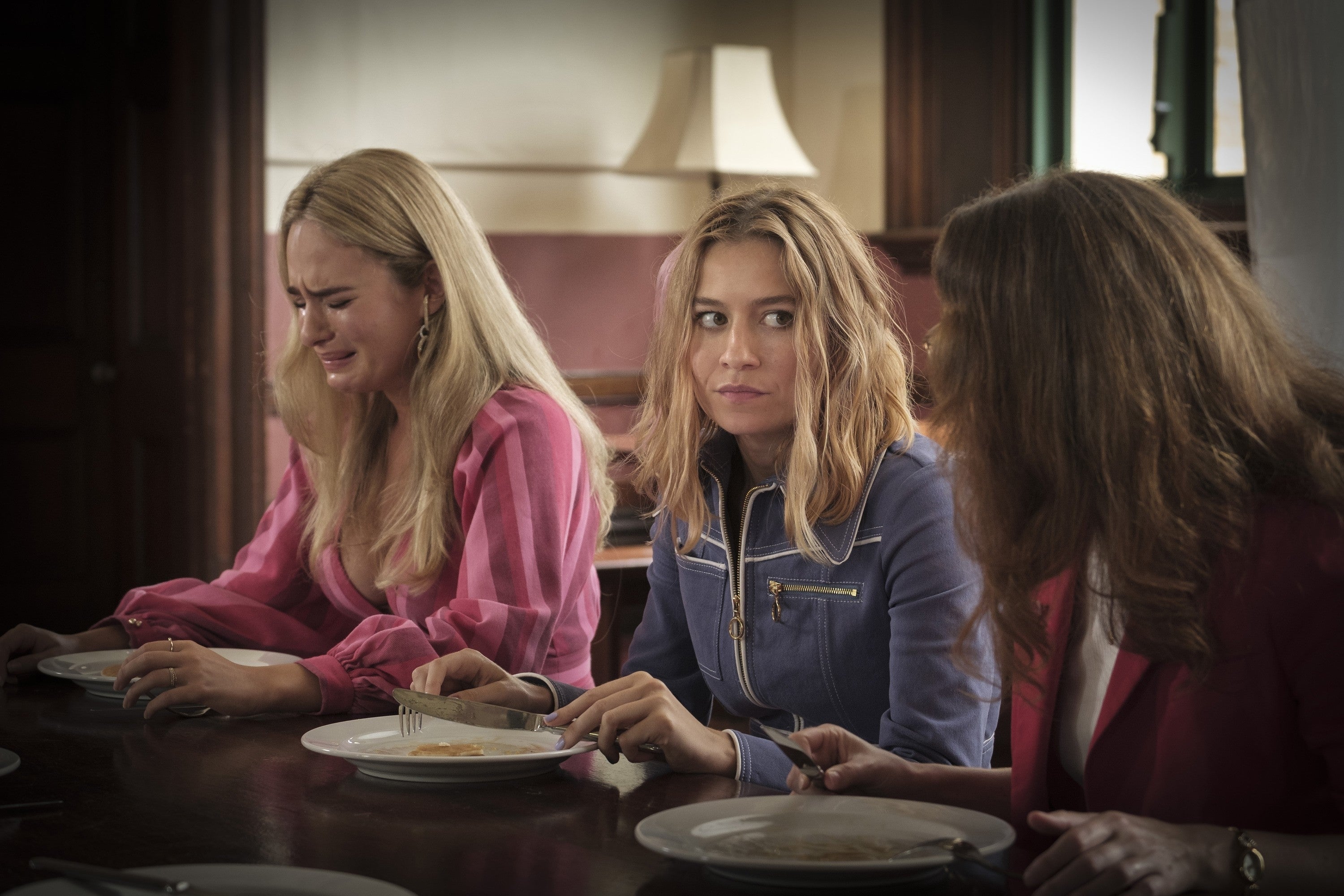 Three women sit at a dining table with empty plates. The woman on the left is crying, while the others look concerned and attentive