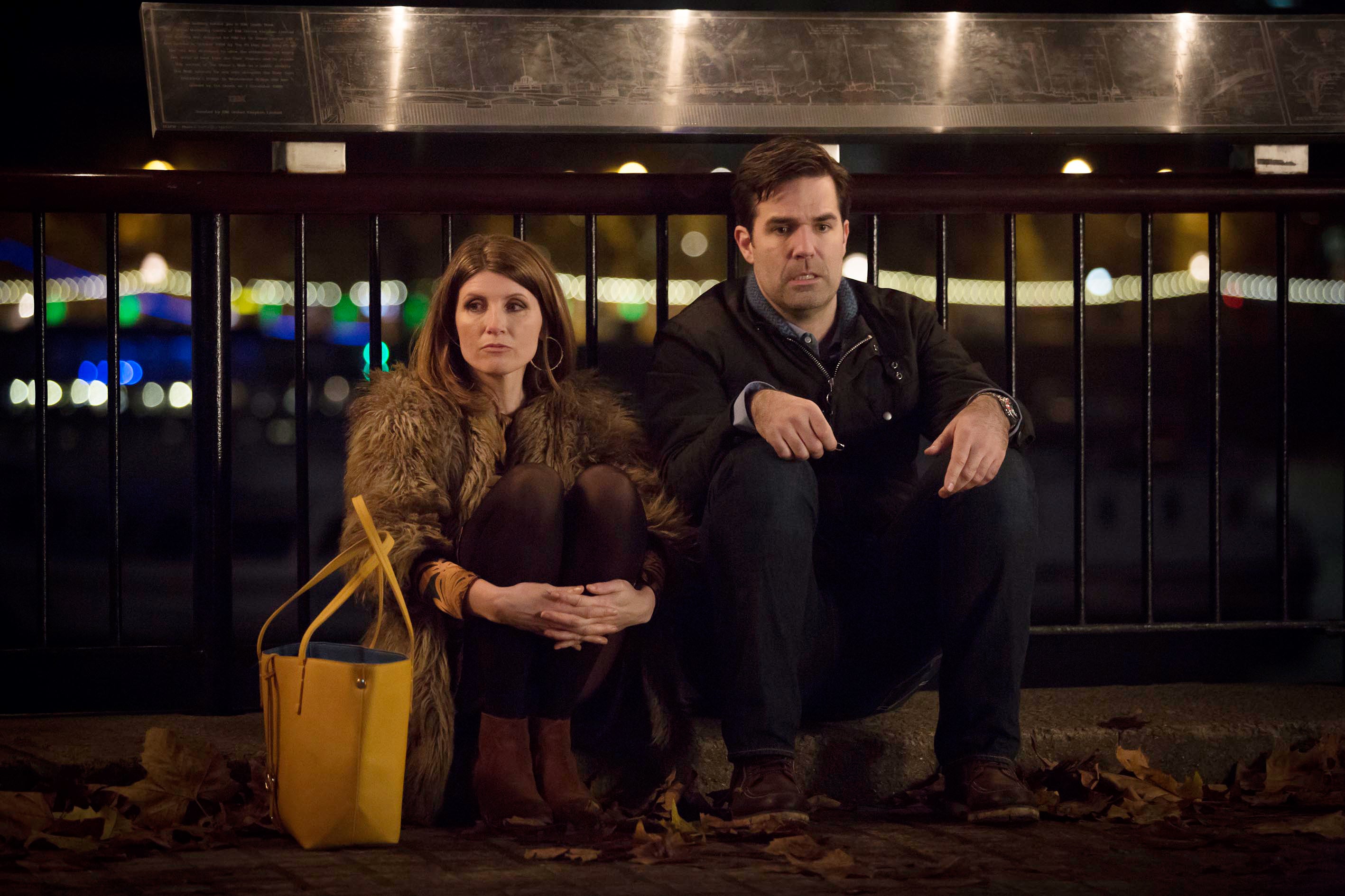 Two people sit by a railing at night; the woman wears a fur coat and has a yellow bag, the man is in a dark coat, both appear contemplative