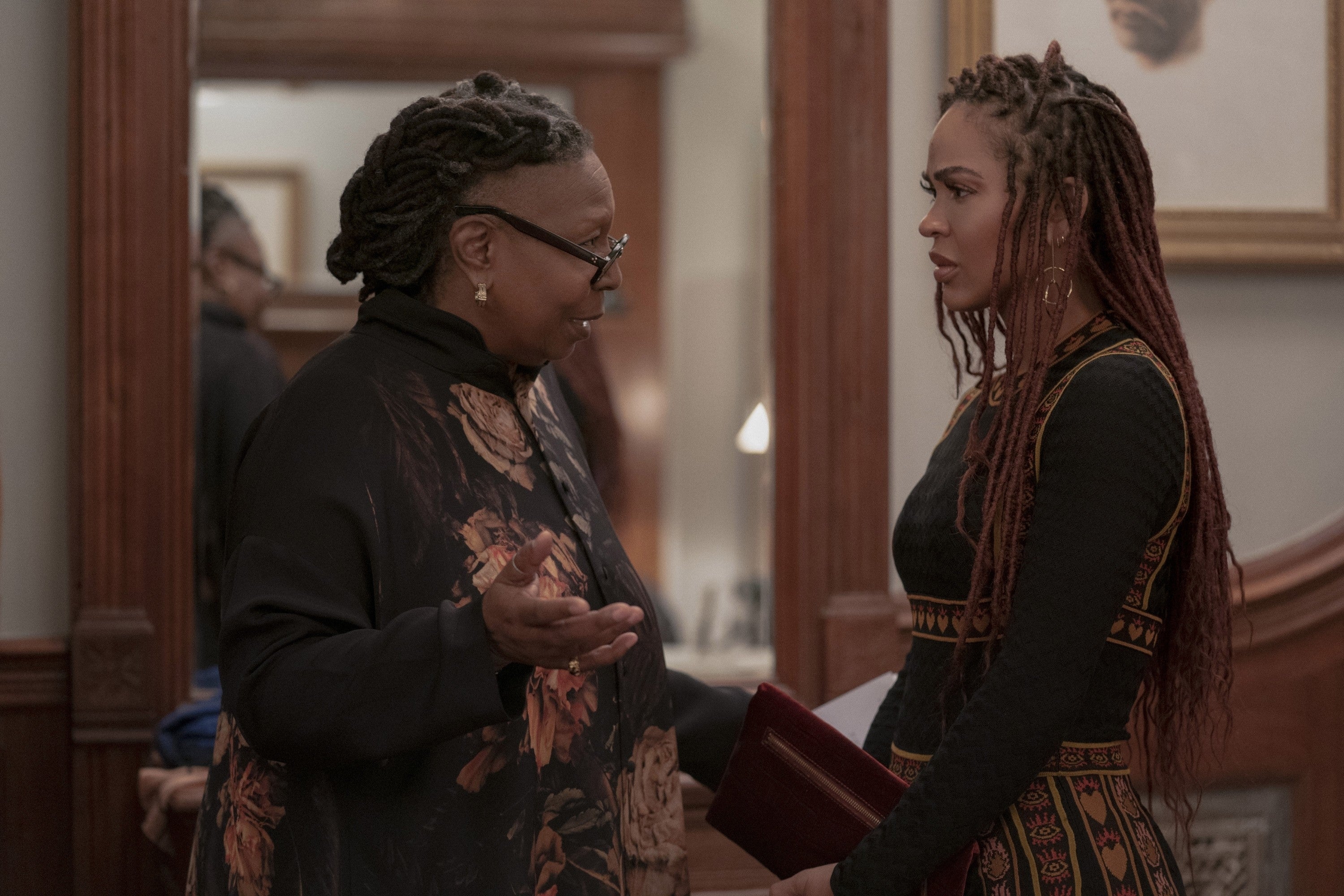 Two individuals in conversation indoors, one with floral outfit and glasses, the other with braided hair and patterned dress, appearing serious