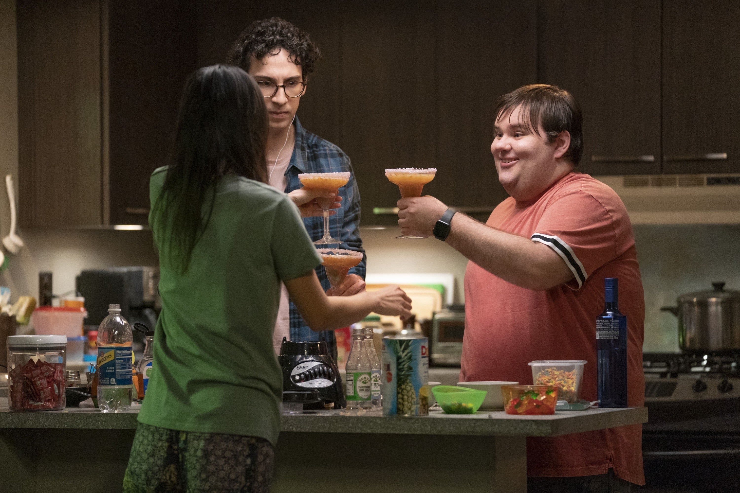 Three people stand in a kitchen, holding drinks and smiling, suggesting a casual, friendly gathering
