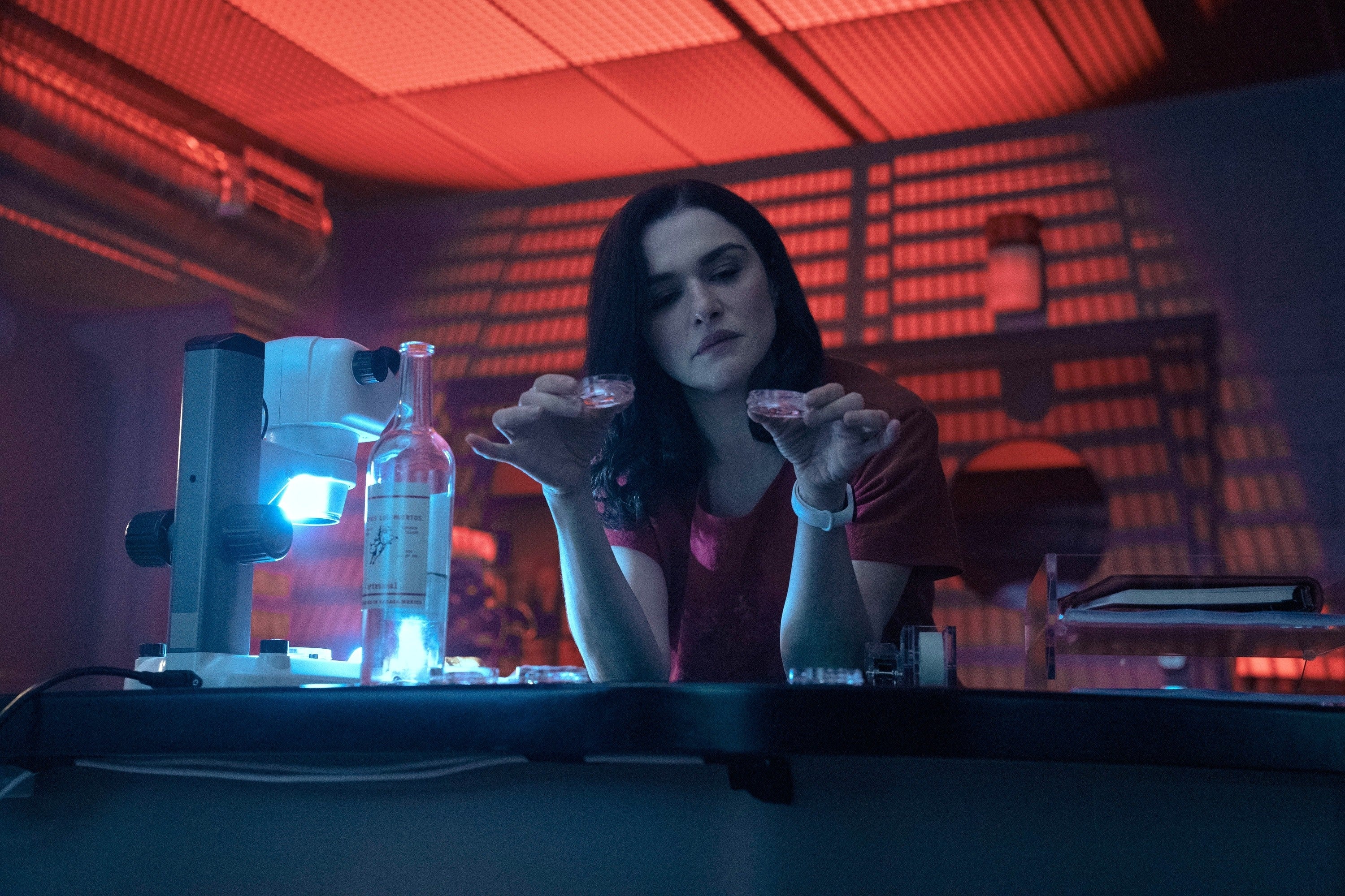 Person in a lab examines samples under a microscope, with a bottle and open notebook nearby, in a dimly lit room