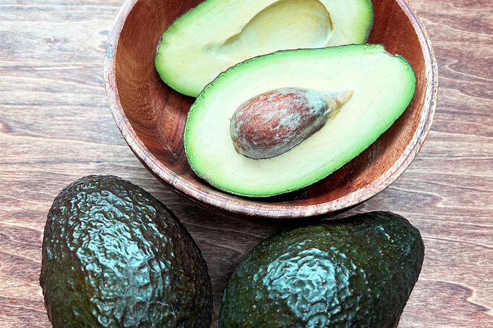 カットされたアボカドが木製のボウルに入り、全体のアボカド2個がその横に置かれています。