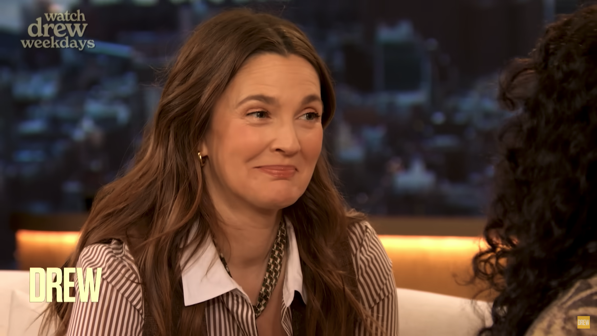 Woman with long hair in a striped shirt smiles during a talk show interview. Background has a cityscape view