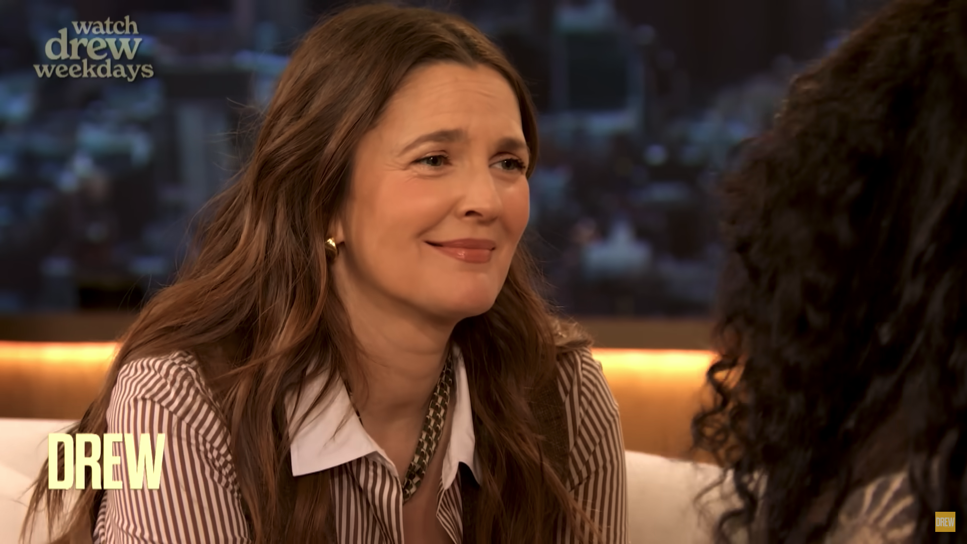 Person smiling during a talk show interview, wearing a striped shirt with a white collar. Background has cityscape visuals