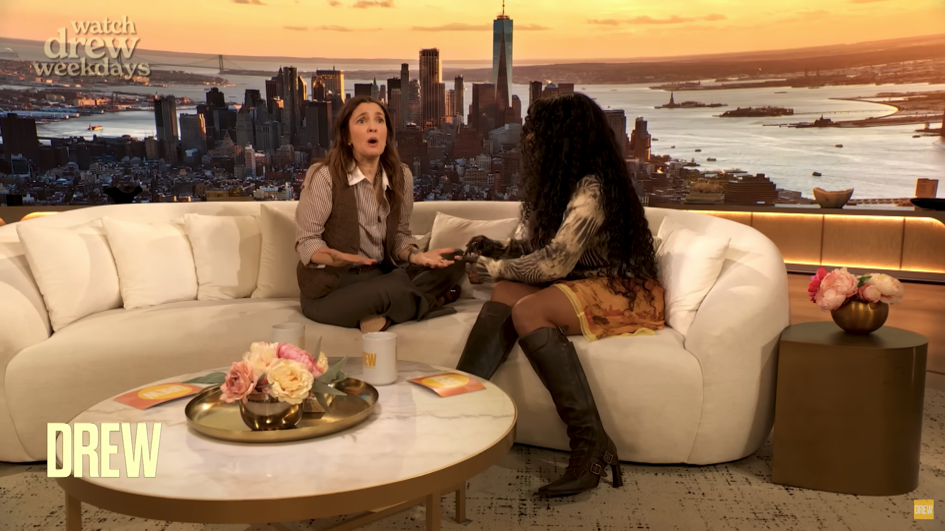SZA and Drew Barrymore sit on a talk show set with a city skyline view. One wears a suit; the other, a floral skirt and boots