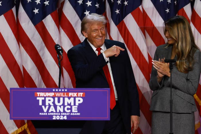 Person in a suit and red tie gestures while standing at a podium with a campaign sign. Flags are displayed in the background