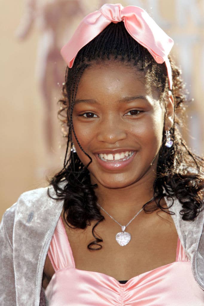 Young person smiling on the red carpet, wearing a satin dress with a matching hair ribbon, earrings, and a necklace
