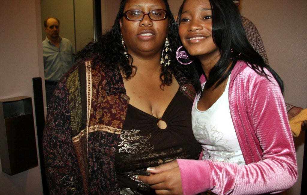 Two people pose together indoors. The person on the right wears a pink jacket over a white top; the person on the left wears a patterned jacket