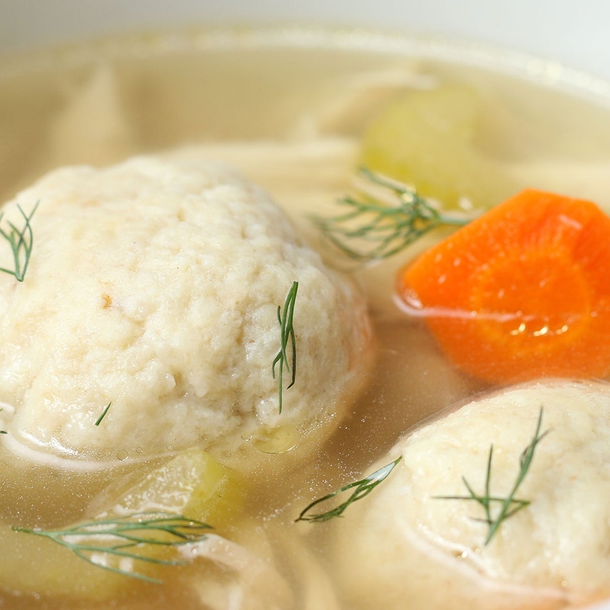 Closeup of matzo ball soup with dill, showcasing fluffy matzo balls, sliced carrots, and celery in broth