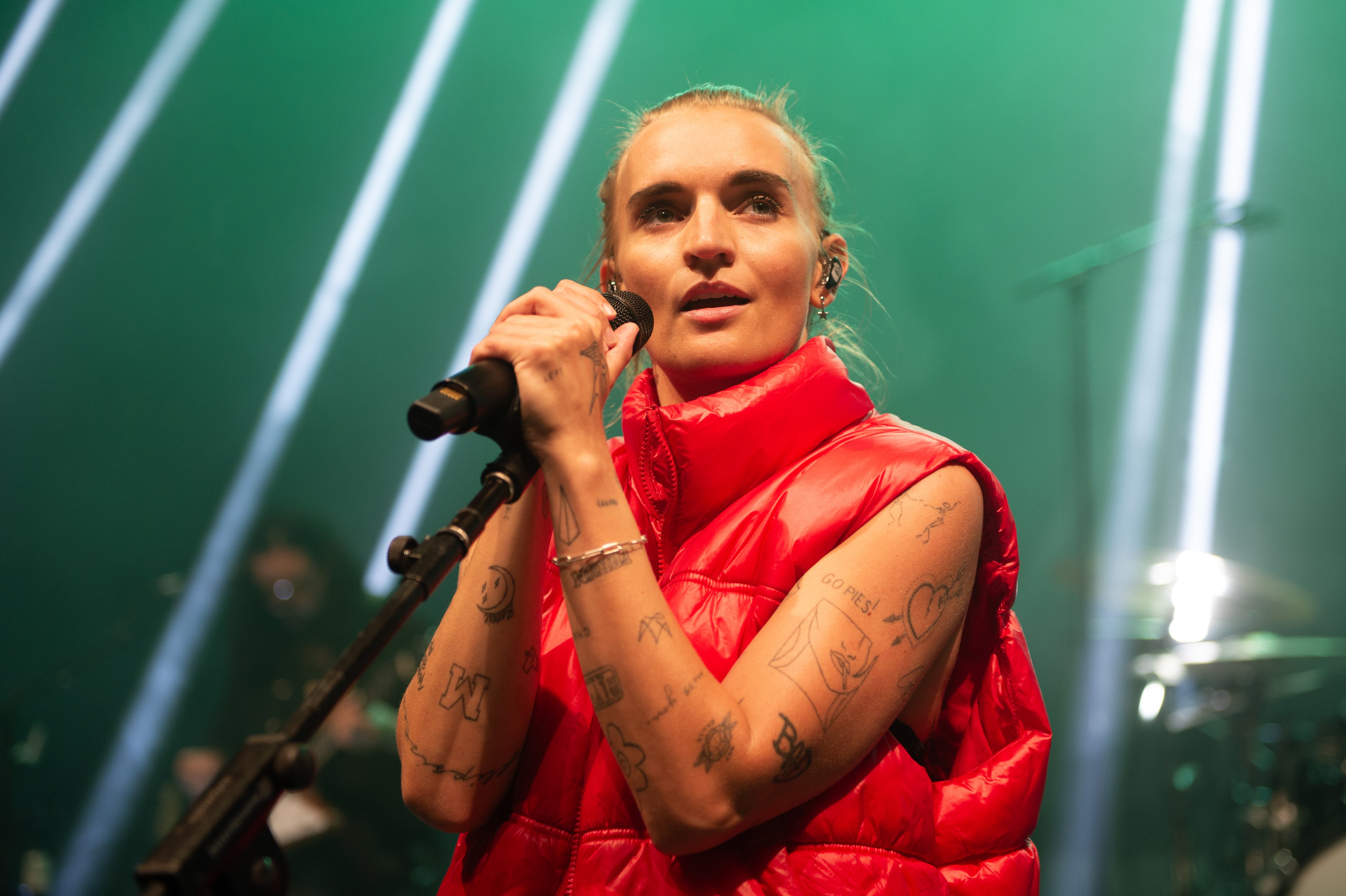 Person on stage with a microphone, wearing a puffy vest, and having visible tattoos on their arms. Stage lights form beams in the background