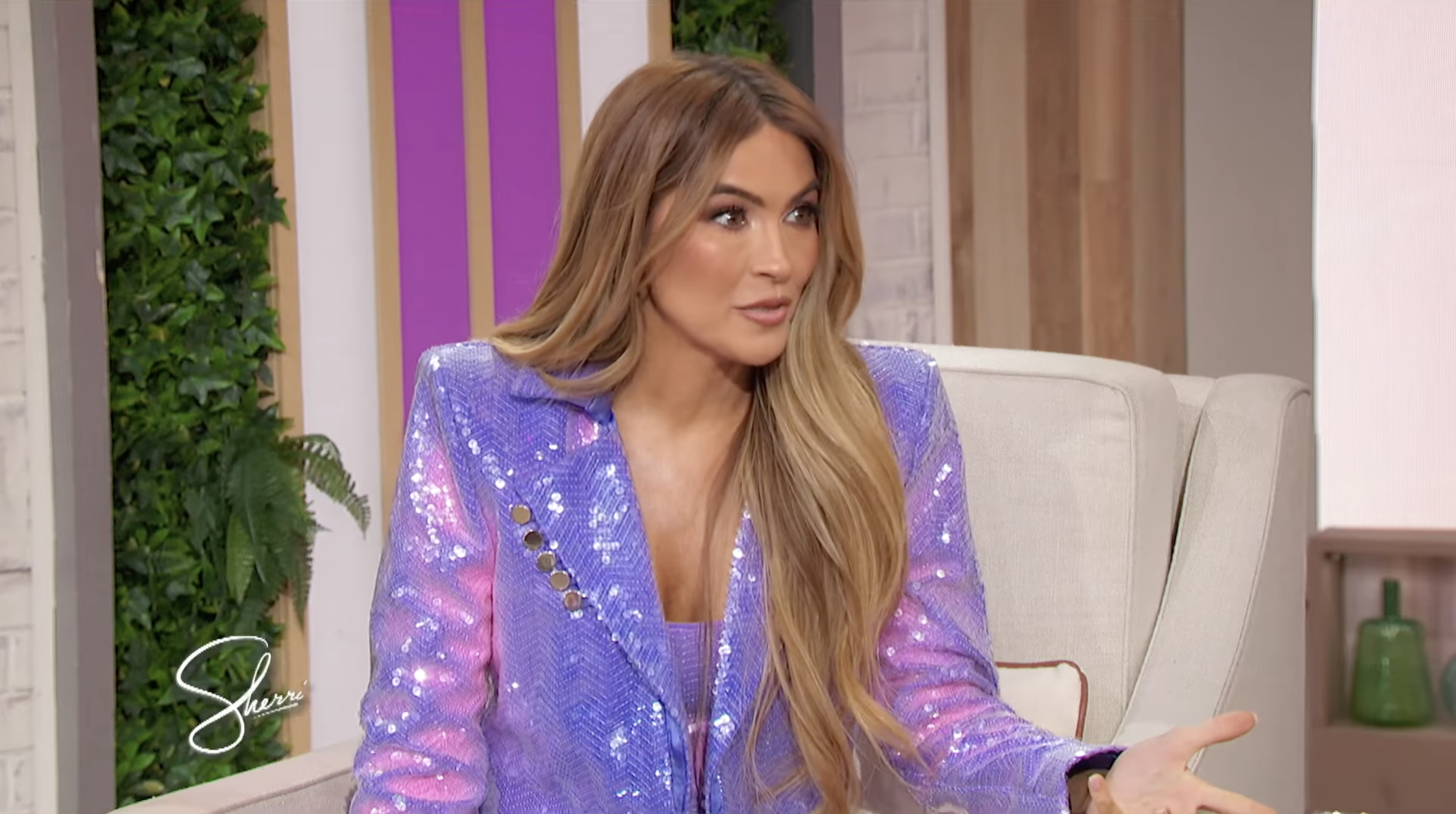 Person in a sparkly, tailored suit on a talk show set, gesturing while speaking