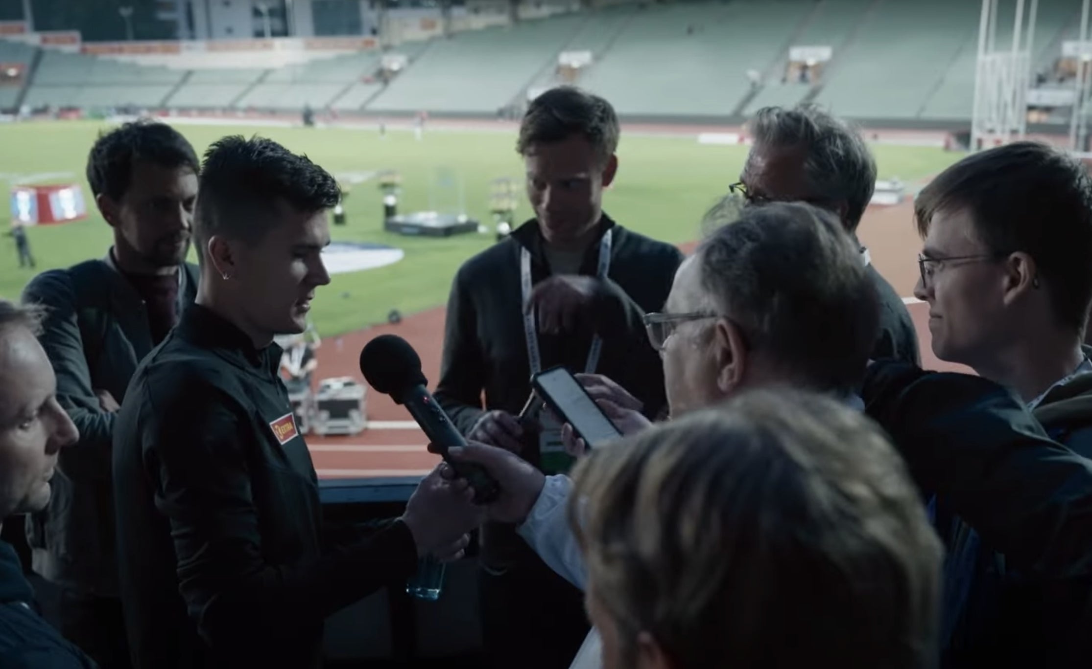 Athlete surrounded by reporters holding microphones and notepads at a sports stadium during an interview