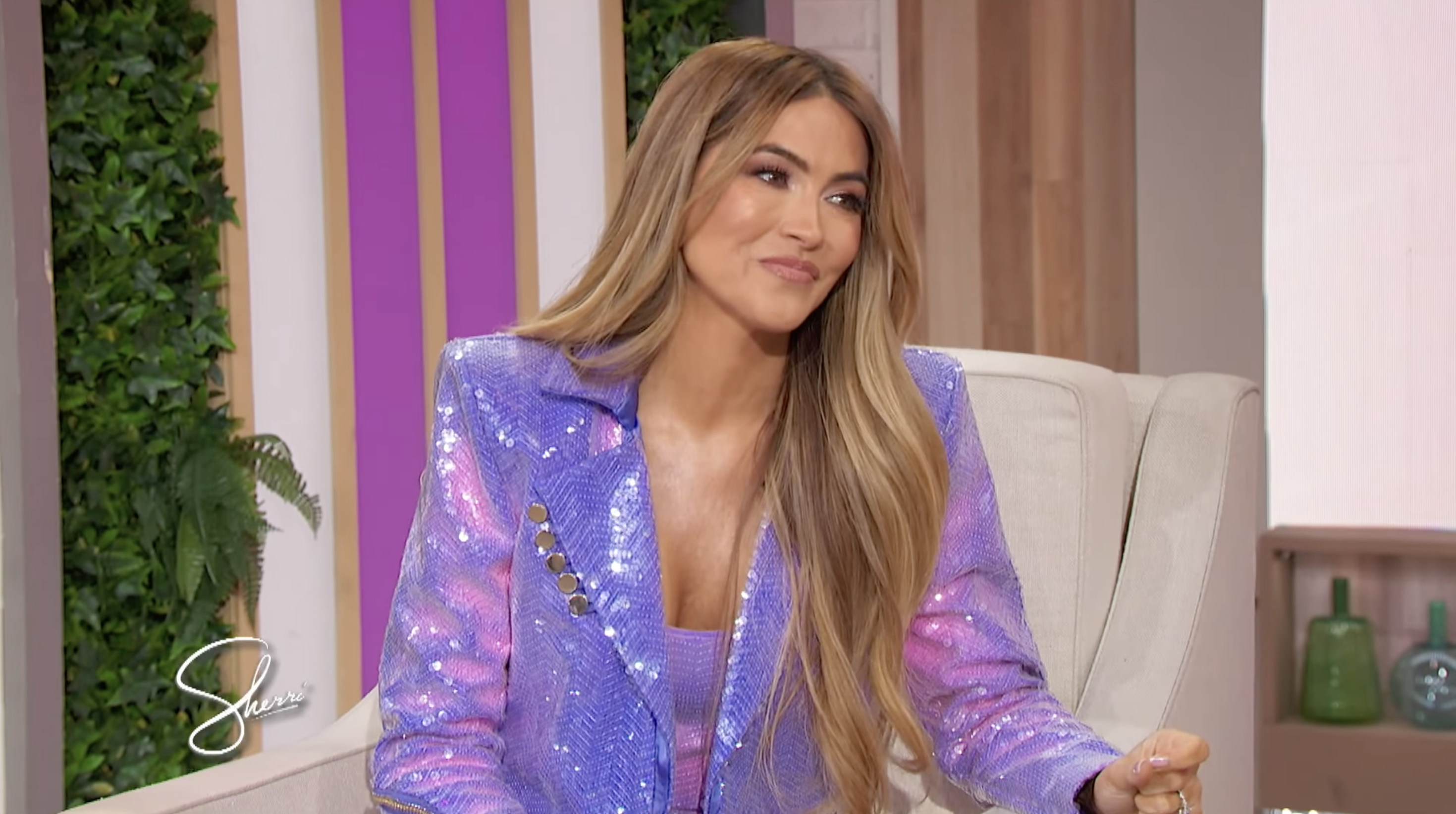 Person seated in an interview setting wears a sparkling blazer with long hair styled down, sitting in a bright studio with plants in the background