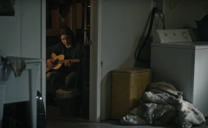 Person sits in a dimly lit room playing a guitar, visible through a doorway. Nearby, laundry appliances and a clothes pile are seen