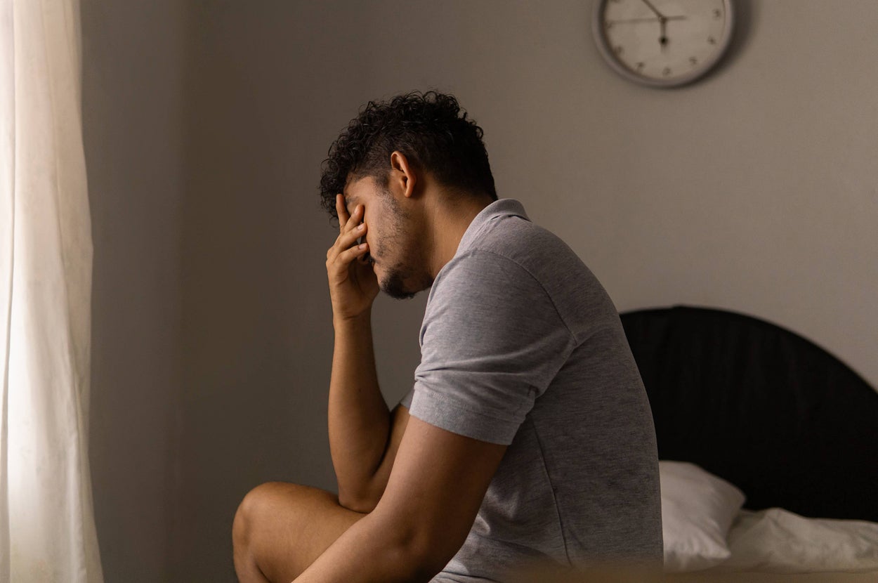 Person sitting on a bed with their hand covering their face, appearing thoughtful or stressed, next to a wall clock showing time