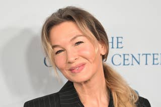 Renée Zellweger smiling at an event, wearing a pinstripe suit with a ponytail hairstyle