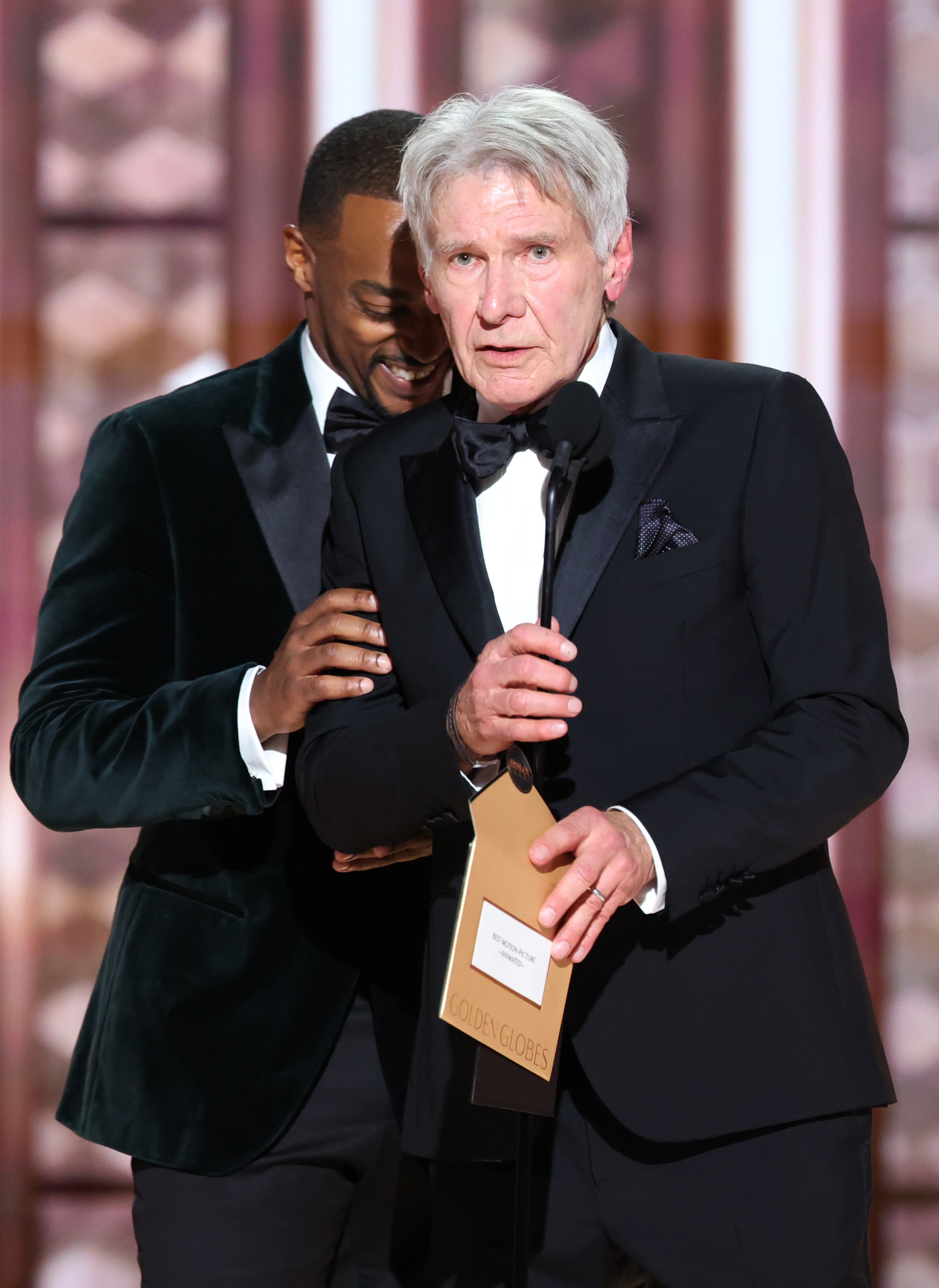 Anthony Mackie and Harrison Ford in tuxedos at an award ceremony, one holding an envelope and speaking into a microphone