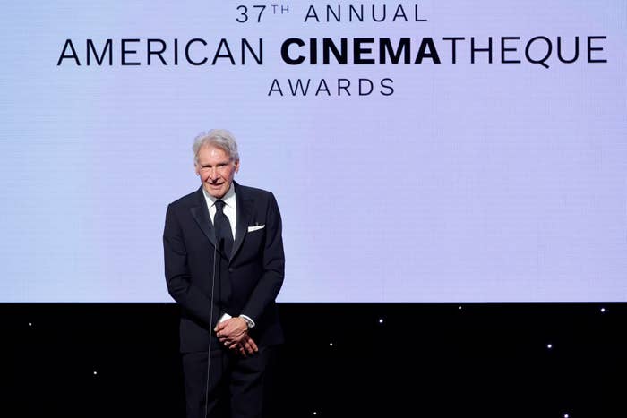 Harrison Ford in suit speaks at the 37th Annual American Cinematheque Awards podium. Background shows award event signage