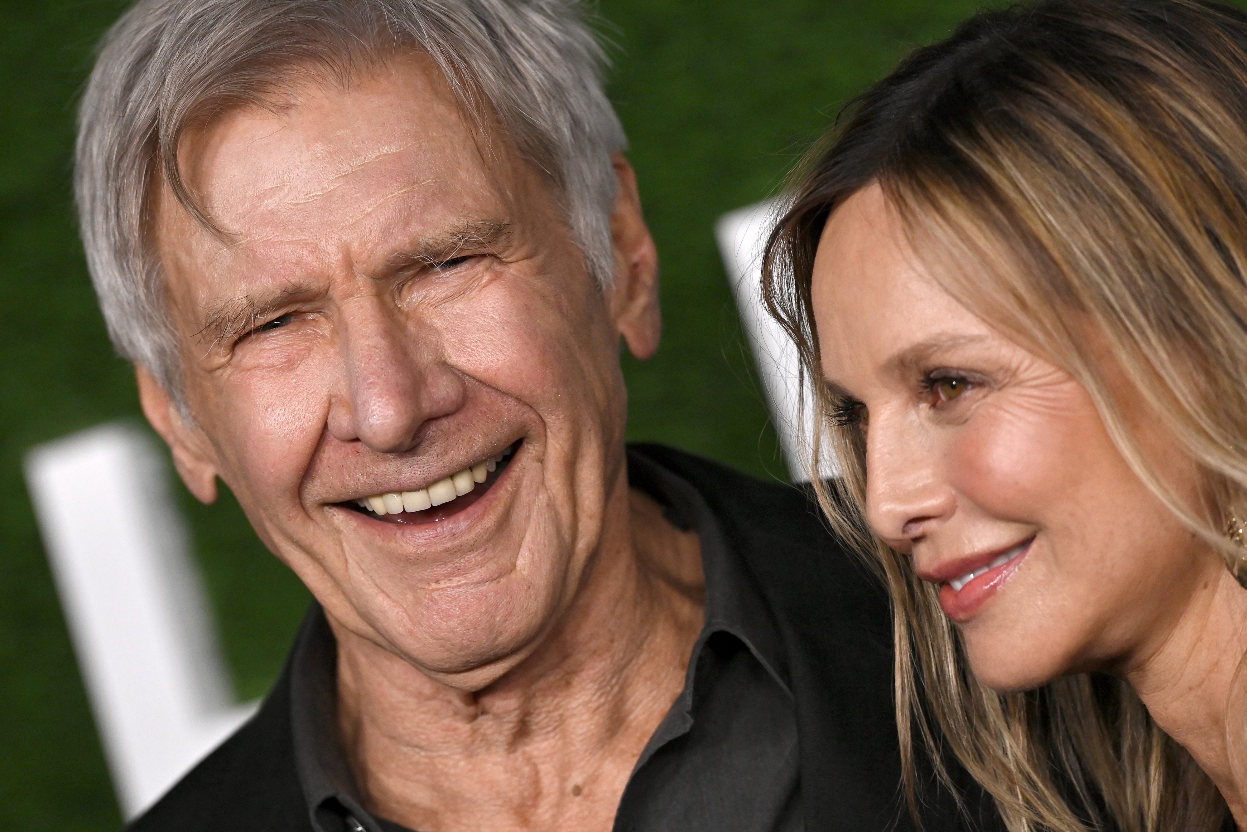 Harrison Ford and his wife smiling at an event. The man is wearing a suit jacket and shirt, while the woman has long hair and appears joyful