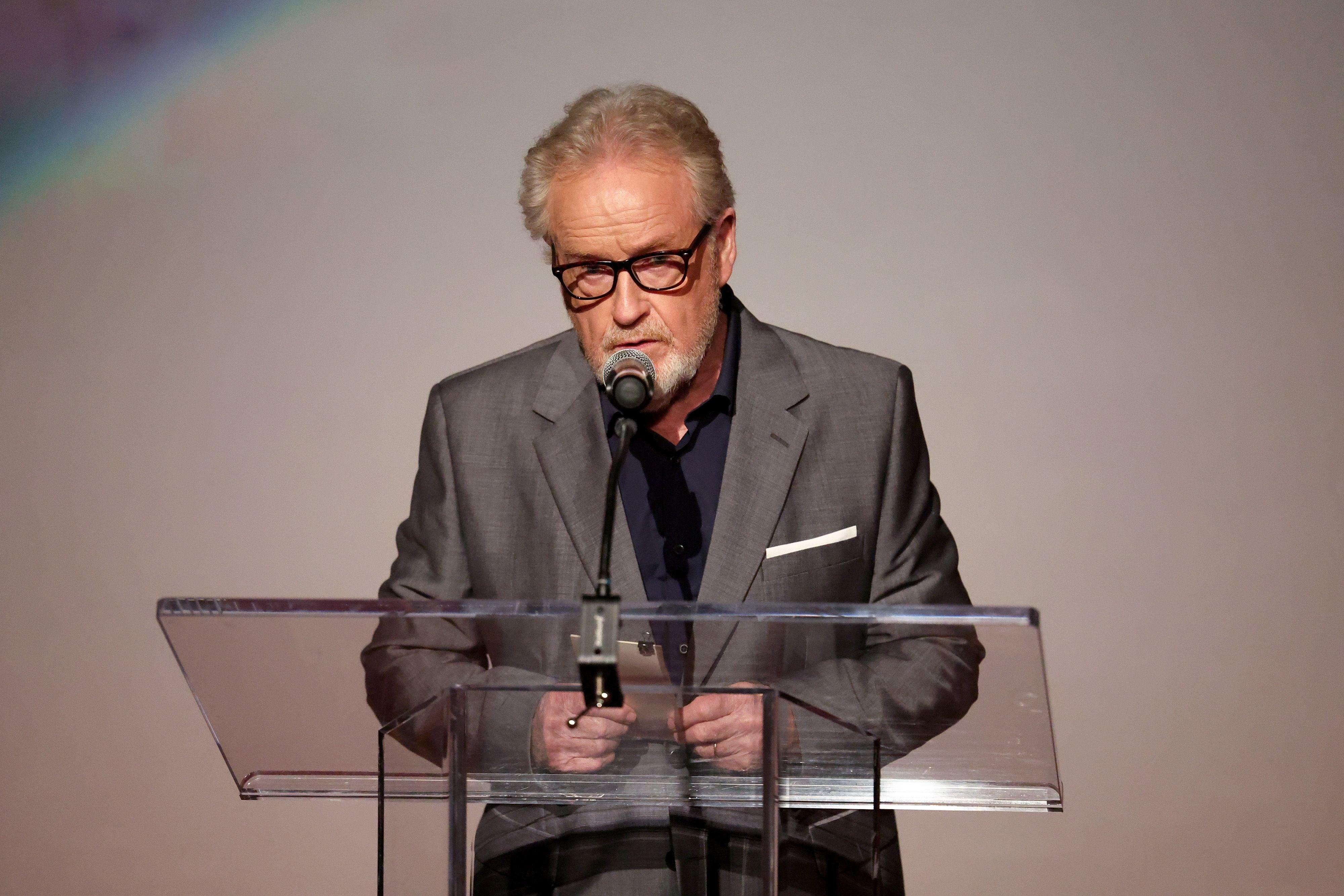 Ridley Scott in a suit stands at a podium speaking into a microphone during an event