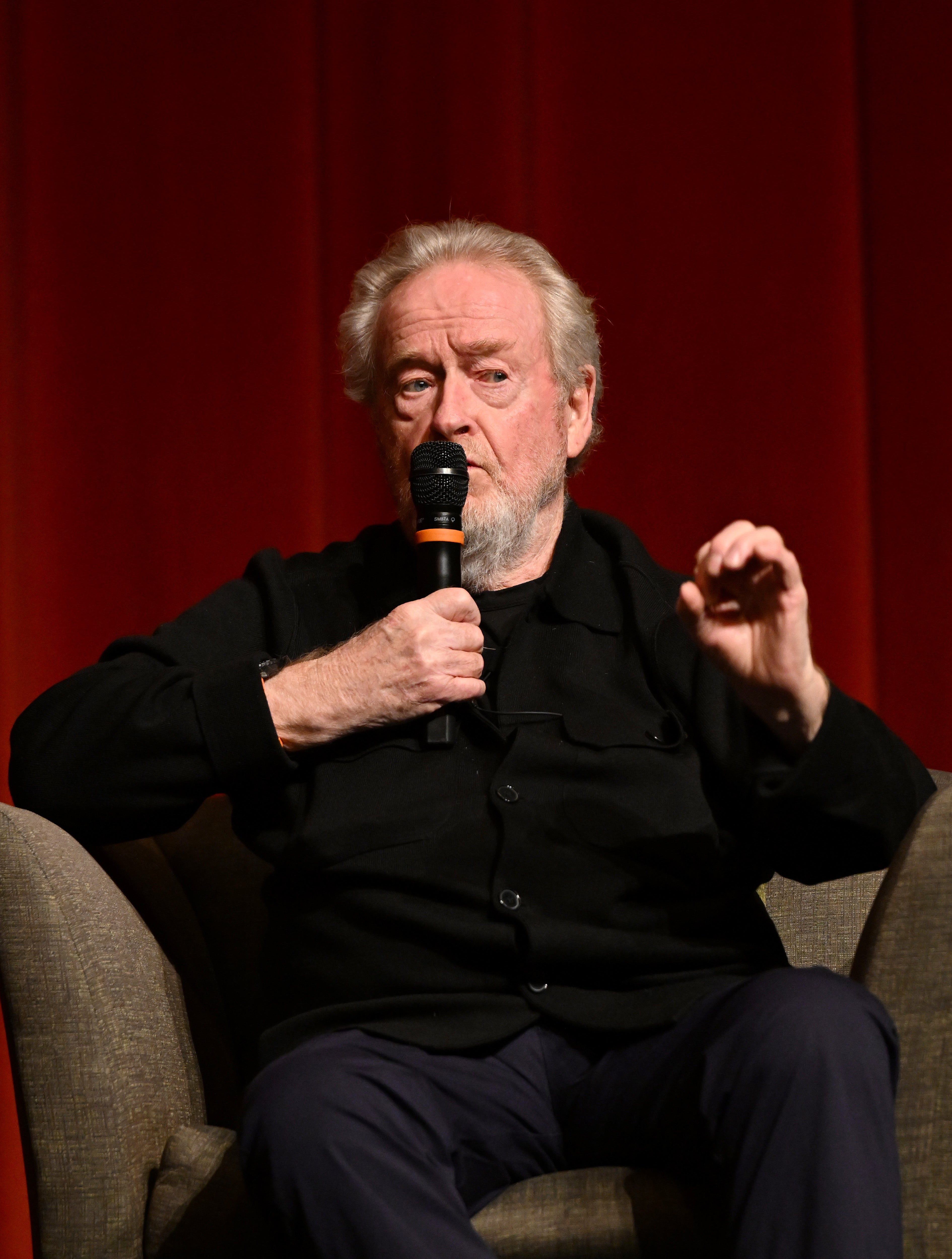 Ridley Scott speaking into a microphone while seated on a stage, wearing a black jacket and dark pants