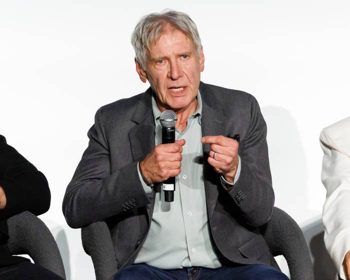 Harrison Ford seated, speaking into a microphone during a panel discussion, wearing a blazer and shirt