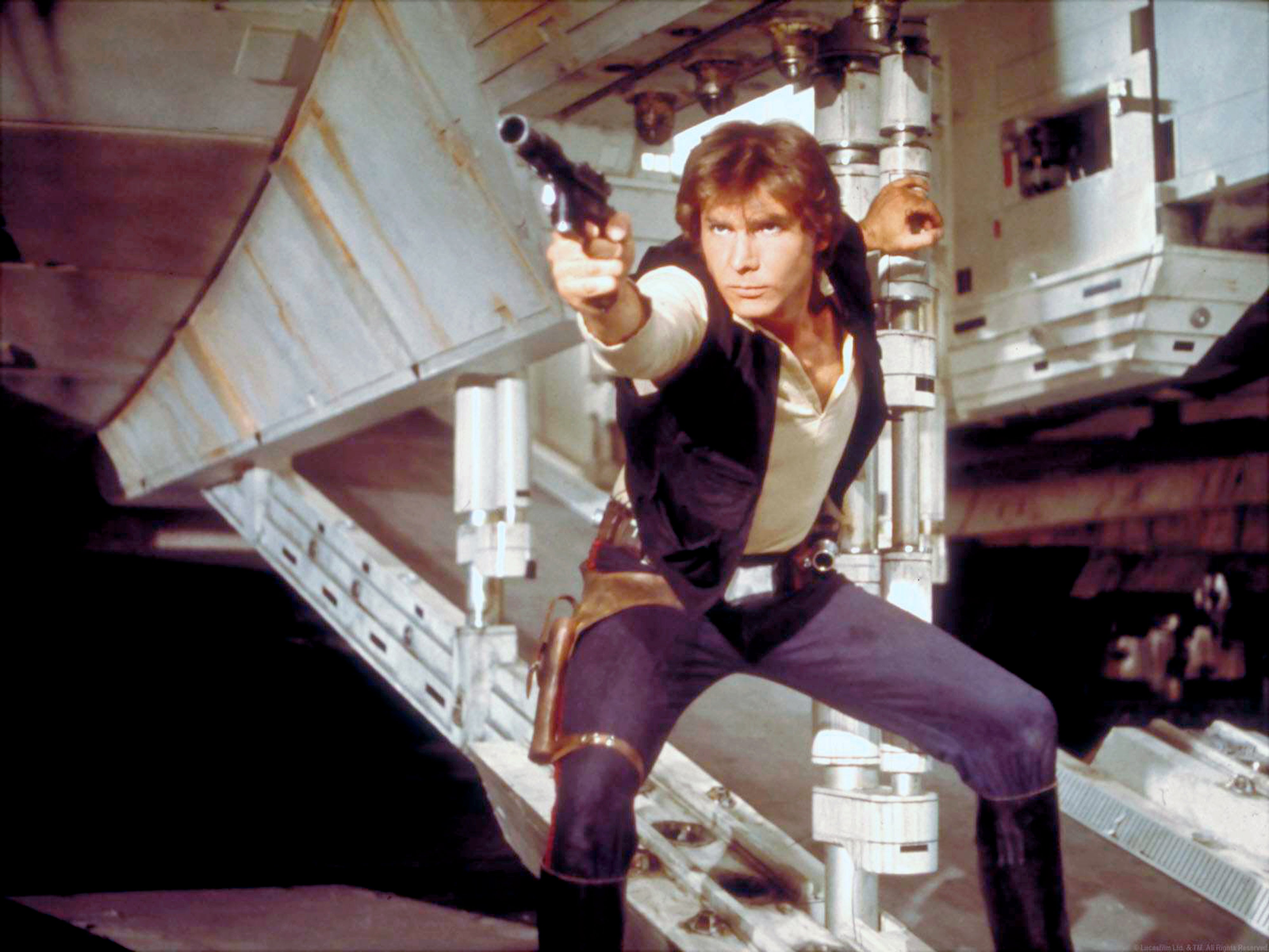 Harrison Ford as Han Solo holds a blaster, posed action-ready on a spaceship set