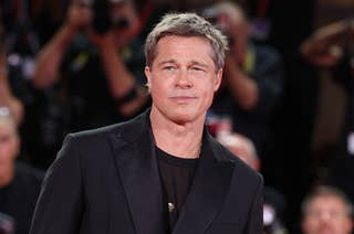 Person wearing a black suit on a red carpet with photographers in the background