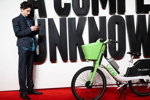 Celebrity in a tailored suit stands next to a Lime bike, using a phone, on a red carpet. Large text backdrop partially visible