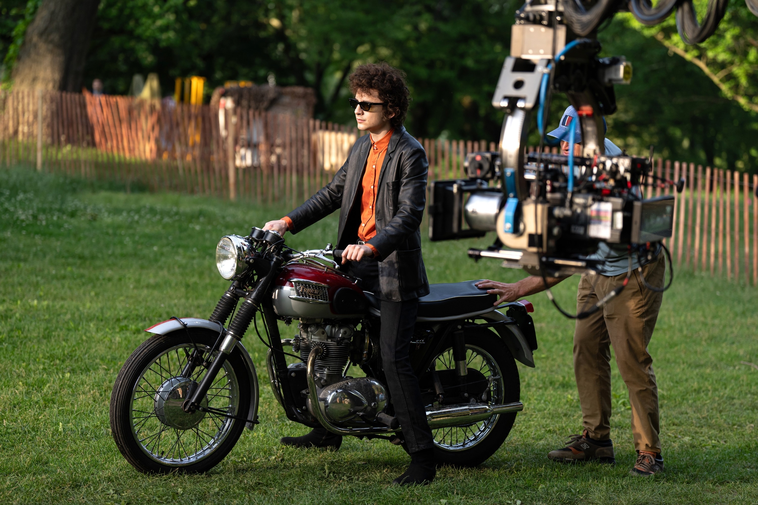 Person in sunglasses and a leather jacket sits on a vintage motorcycle during a film shoot outdoors