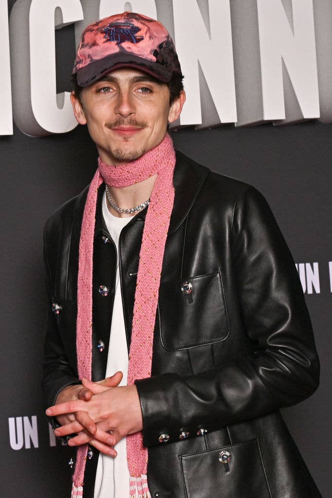 Person wearing a stylish leather jacket and pink scarf on a red carpet, in front of a backdrop with large text