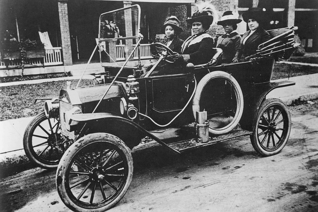 Four women dressed in early 20th-century attire sit in a vintage car, parked on a residential street