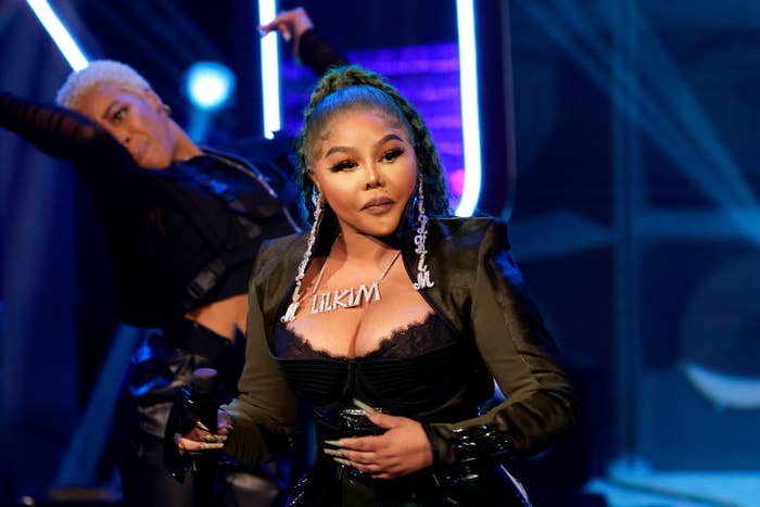 Person performing on stage in stylish outfit with bold accessories, holding a microphone. Background dancer is visible