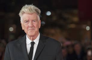 David Lynch in a suit and tie stands on a red carpet, with a blurred background of people and lights