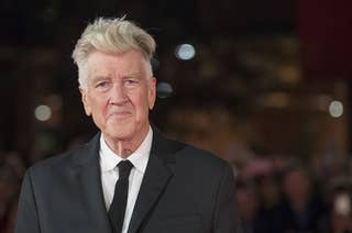David Lynch in a suit and tie stands on a red carpet, with a blurred background of people and lights