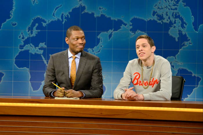 Two people are seated at a news desk on a set with a world map background. One is in a suit, the other in a casual sweatshirt