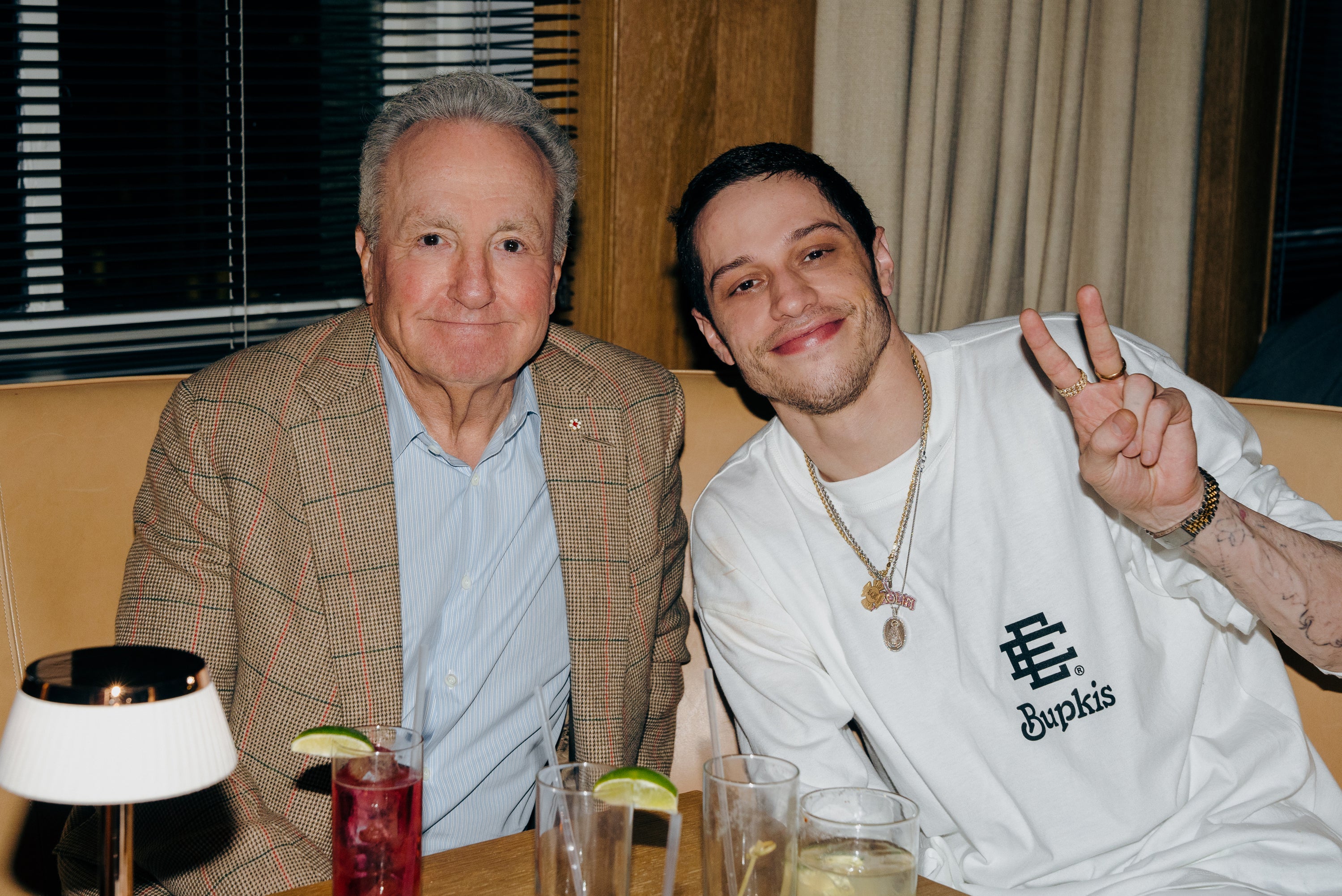 Two people seated together; one in a checked blazer, the other in a casual T-shirt, making a peace sign. Glasses with drinks on the table