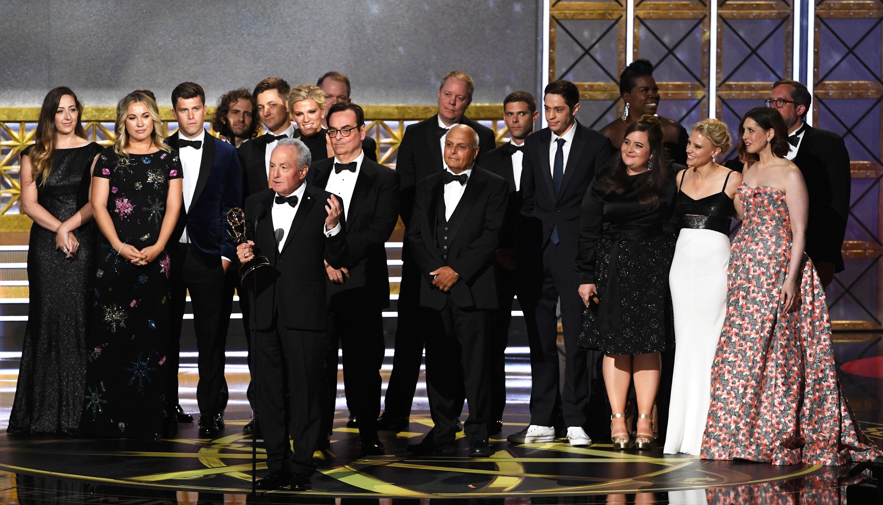Group of celebrities in formal attire at an awards ceremony, some holding trophies on stage