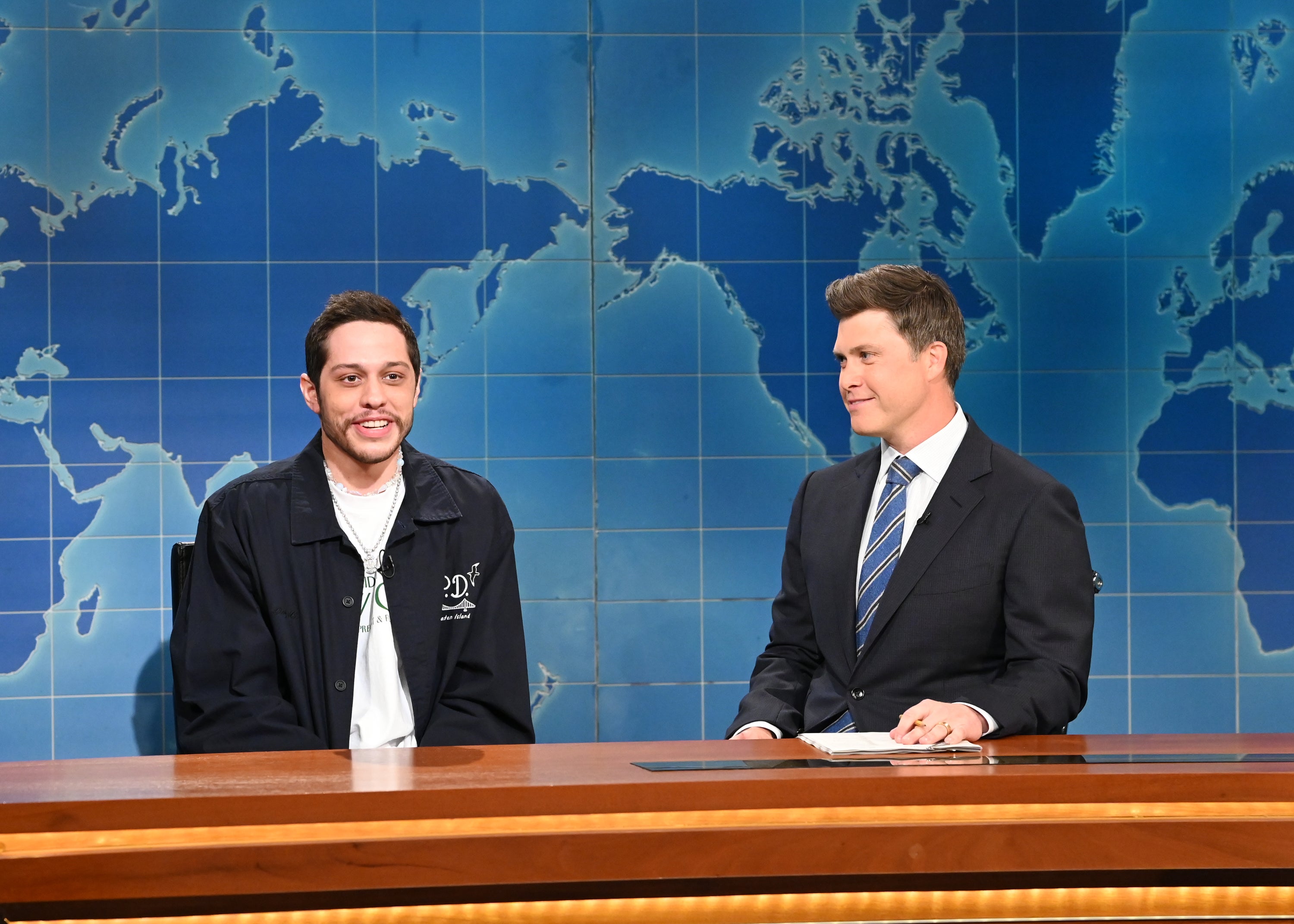 Two men sitting at a news desk on a TV set. One wears casual attire with a jacket; the other is in a suit and tie. A world map is in the background
