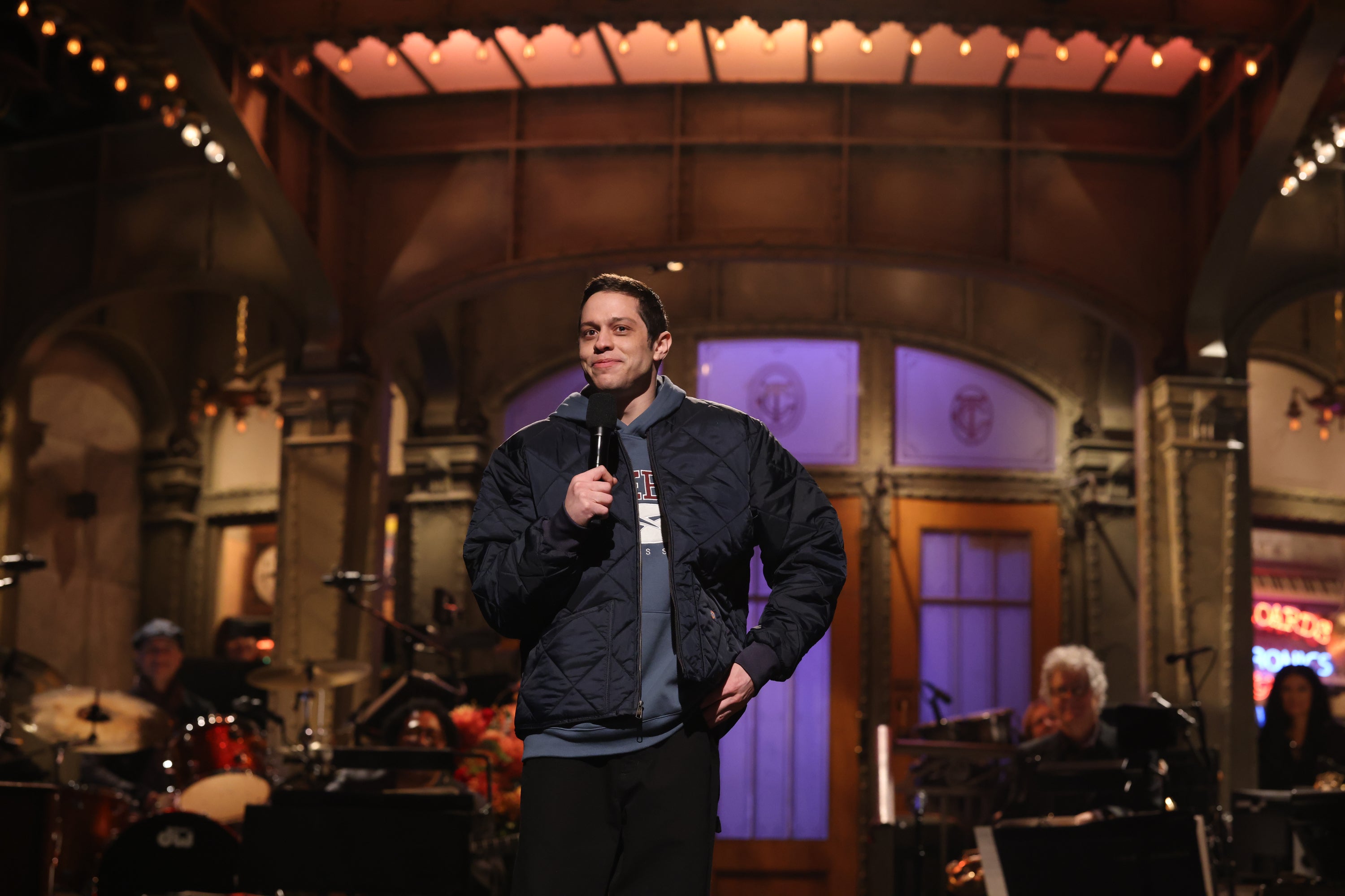 Comedian holding a mic on a stage with a band in the background, wearing a casual quilted jacket and sweatshirt