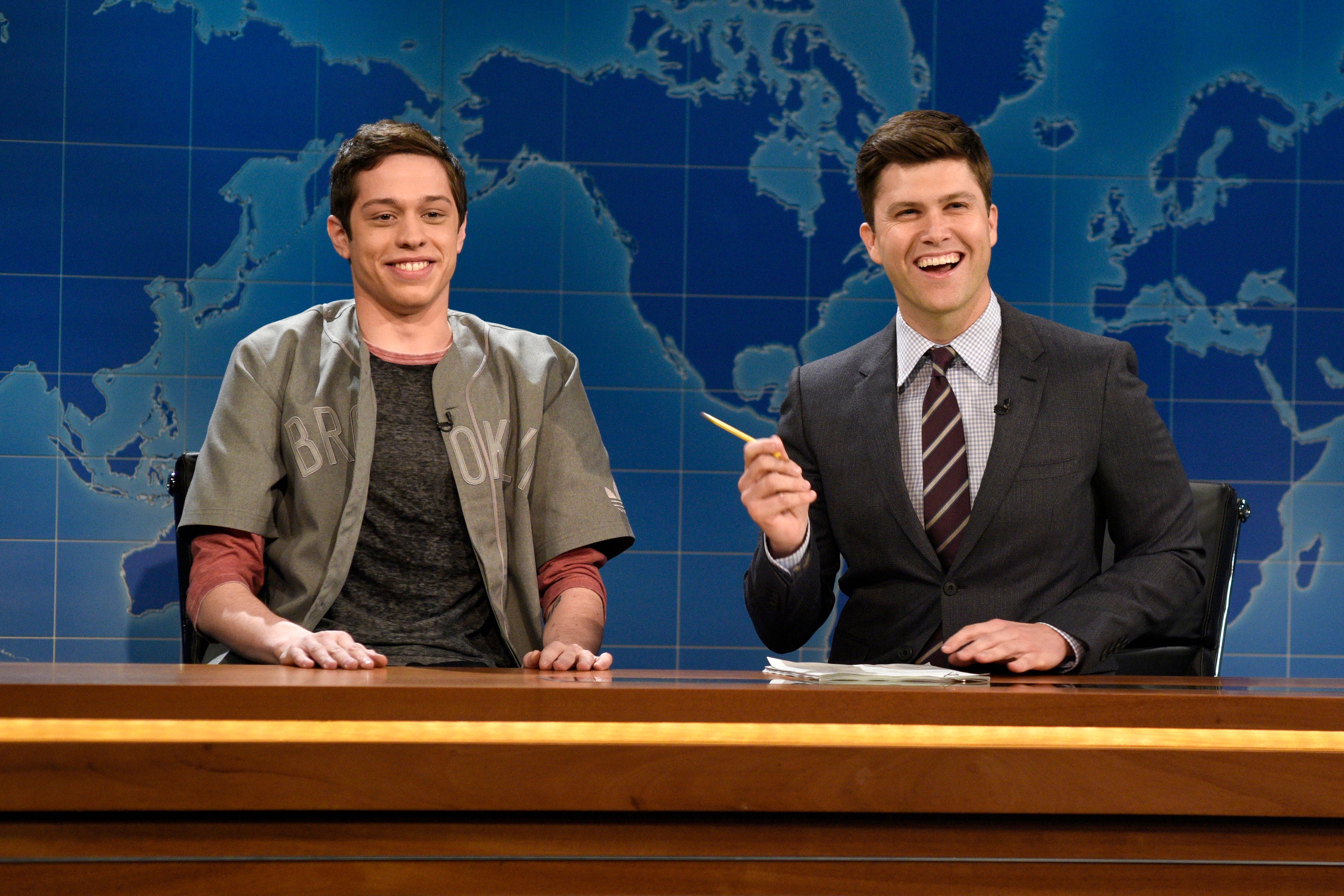 Two people sit behind a news desk. One wears a casual shirt, the other a suit with a tie, both smiling brightly
