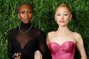 Ariana Grande and Cynthia Erivo in elegant dresses on a lush green backdrop: Cynthia in black with a sheer top, Ariana in a strapless pink gown, both with jewelry