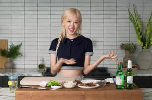 Person with braided hair stands behind a kitchen counter with ingredients and drinks, smiling and gesturing in a cooking demonstration