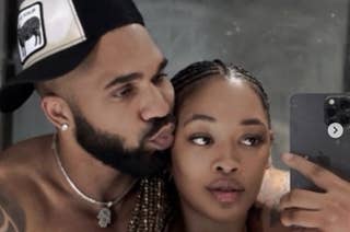 Two individuals taking a mirror selfie, one with a cap and beard, playfully kissing the other's cheek. They appear casually dressed