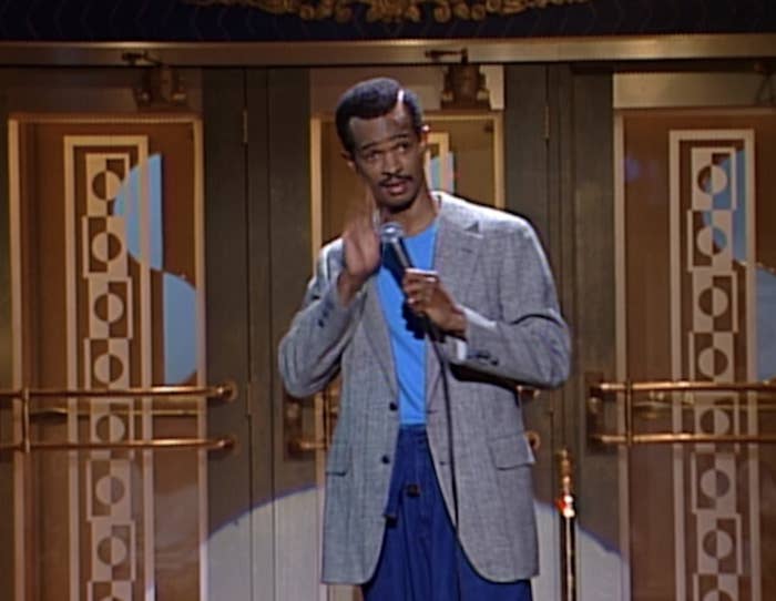Person on stage performing stand-up comedy, wearing a gray blazer and blue shirt, holding a microphone. Art Deco style doors in the background