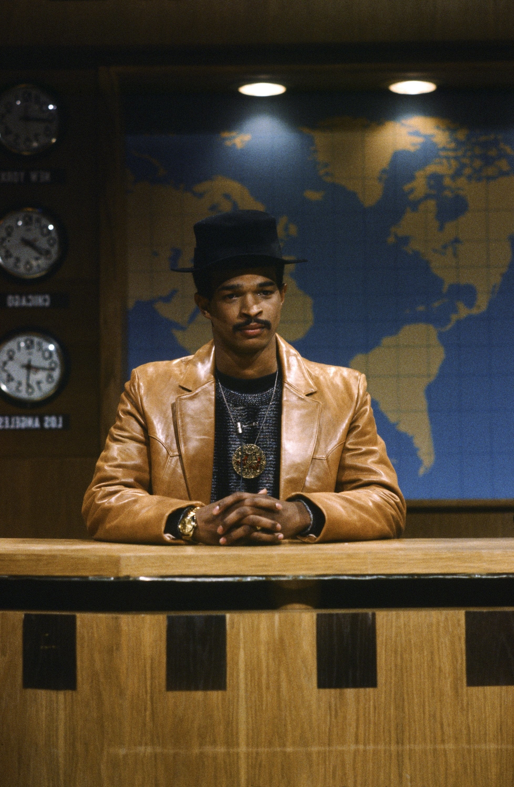 A person sits at a news desk wearing a fedora and leather jacket, in front of a world map